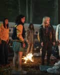Group of people standing around a campfire in a forest, with a crashed plane in the background.