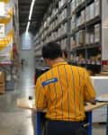 Store employee in yellow striped shirt working in a large warehouse aisle with shelves of boxed furniture.