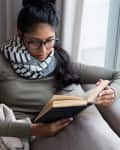 Woman in glasses reading a book on a beige sofa, wrapped in a blanket, wearing a striped scarf.