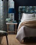 Bedroom with floral upholstered bed, blue lamp, and floral room divider. Beige knit throw on white bedding.