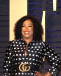 Person in a black and white polka dot dress with a gold belt, standing in front of a dark blue backdrop.