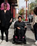 Three people walking on a city sidewalk, one using a wheelchair, with red and black storefronts in the background.