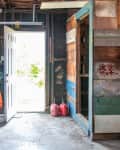 Cluttered garage with open door, exposed beams, shelves, tools, and two red gas cans on the floor.
