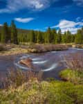 Mountain stream flowing through a forested landscape with clear blue sky and distant snow-capped peaks.