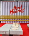 Outdoor seating area with yellow bench, concrete table, and Jenga blocks on red carpet, striped backdrop with "Hôtel Hennes."