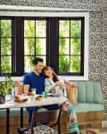 Couple sitting on a teal banquette in a floral wallpapered room, with a table set for breakfast.