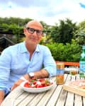 Man in blue shirt sitting at outdoor table with salad, charcuterie, and drinks, surrounded by greenery.