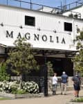 Exterior of Magnolia building with large sign, surrounded by trees and flowers, and three people walking towards the entrance.