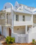 White stucco and wood beach house with balconies, arched windows, and a gated entrance, surrounded by palm plants.
