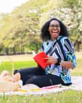 Woman sitting on a picnic blanket in a park, holding a red book and smiling, with a basket nearby.
