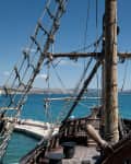 View from a ship's deck with rigging and steering wheel, overlooking a blue sea and distant mountains.