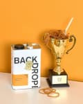 Paint can labeled "Backdrop" next to a gold trophy filled with rubber bands on an orange background.