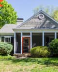 Single-story house with gray siding, orange door, and screened porch, surrounded by lush green bushes and trees.