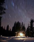 Cabin in snowy forest under starry night sky with visible star trails.