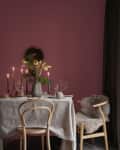 Dining table with white tablecloth, candles, floral centerpiece, and wooden chairs against a mauve wall.