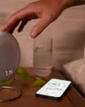 Hand reaching for a round digital clock on a bedside table with a smartphone, glass of water, and green plant.