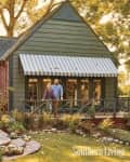 Couple standing on porch of green house with striped awning, surrounded by autumn foliage and pumpkins.