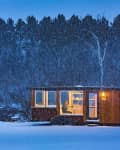 Tiny house with warm interior lighting in snowy forest setting at dusk.