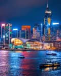 Vibrant city skyline of Hong Kong at night with illuminated skyscrapers, a modern building, and boats on the water.