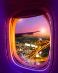 Cityscape view from an airplane window at sunset, with a wing and illuminated streets below.