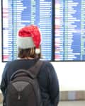 A person wearing a Santa hat stands in front of a flight information display board at an airport.