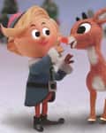 A cheerful elf in a blue outfit interacts with a red-nosed reindeer in a snowy landscape.