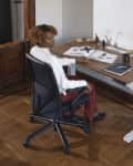 Person sitting at a wooden desk with books, a lamp, and a plant, in a room with wooden flooring and bookshelves.