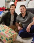 Two men smiling while sitting on a carpet in a festive living room, with a large wrapped gift and holiday decorations.