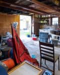 Cluttered garage with wooden walls, storage bins, folding chair, and various tools. Open door leads to outdoor area.