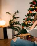 Woman reading a book in a cozy chair next to a decorated Christmas tree with red and gold ornaments.