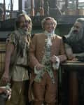 Three men in period costumes on a ship, one in a tan suit, another in rugged attire, and the third with long gray hair.