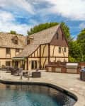 Tudor-style house with a pool, patio furniture, outdoor kitchen, and a yellow cabana in a lush garden setting.