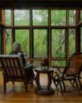 Man sitting in a sunroom with large windows, surrounded by trees, wooden shelves, and a rocking chair.