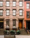 Row of classic brownstone townhouses with black and wooden doors, iron railings, and small trees in front.