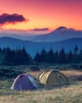 Two tents on a grassy field with a forest and mountains at sunset.