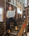 Woman holding blueprints on wooden stairs in a house under renovation.