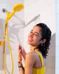 Woman smiling in shower with yellow showerhead, phone holder, and plant on white tiled wall.