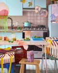 Colorful kitchen with blue cabinets, patterned tablecloths, hanging paper lantern, and fresh produce on the table.