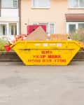Yellow skip bin filled with debris in front of brick houses and flowering bushes.