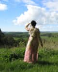 Woman in floral dress standing in grassy field, looking at a scenic landscape with trees and clouds.