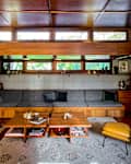 blue cushion built in couch with low hanging wood ceiling detail and rectangle slit windows across top of room, yellow chair and angled fireplace