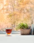Glass mug of tea, wicker basket with green plant, and vintage books on a windowsill with raindrops outside.