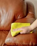 Hand cleaning a brown leather sofa with a yellow cloth.