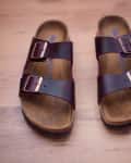 Brown leather sandals with two adjustable straps and buckles on a wooden floor.