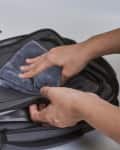 Hands cleaning a gray backpack with a cloth, focusing on the front pocket.