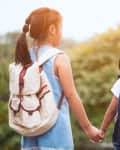 Two children with backpacks holding hands with an adult, standing outdoors.