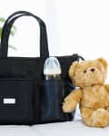 Diaper bag with a baby bottle in pocket, next to a plush teddy bear on a bed.