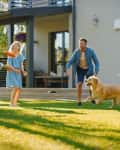 Family playing frisbee with a dog on a sunny lawn in front of a modern house.