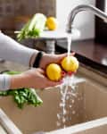 Washing two lemons under a kitchen faucet with celery and a potted plant nearby.