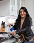 Woman cleaning a kitchen stove with a spray bottle and cloth, next to a teal kettle and knife block.